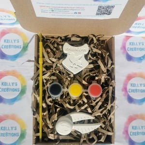 May include: A craft kit in a cardboard box, featuring a Ravenclaw crest, paint pots in black, yellow, and red, a yellow paintbrush, and a Snitch. The kit includes decorative paper filler. The box is surrounded by colorful stickers.