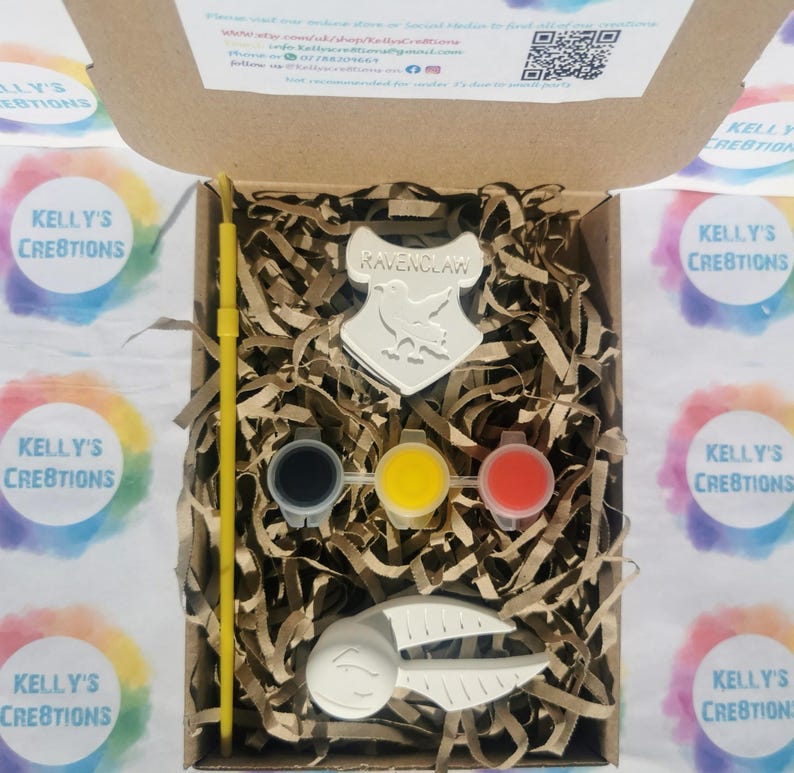 May include: A craft kit in a cardboard box, featuring a Ravenclaw crest, paint pots in black, yellow, and red, a yellow paintbrush, and a Snitch. The kit includes decorative paper filler. The box is surrounded by colorful stickers.