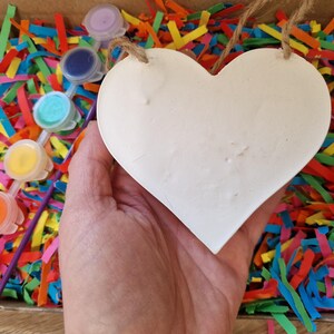 Puede incluir: Un coraz&oacute;n blanco para manualidades con una cuerda de yute, sostenido sobre una caja llena de papel triturado de colores. Se ven peque&ntilde;os botes de pintura roja, naranja, amarilla, turquesa, azul y morada, con pinceles.