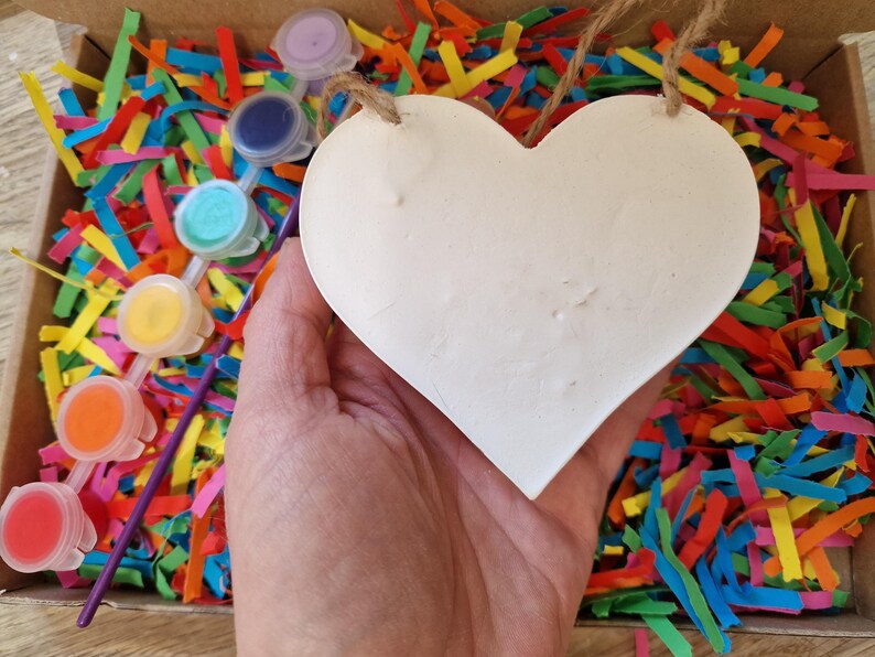 Puede incluir: Un coraz&oacute;n blanco para manualidades con una cuerda de yute, sostenido sobre una caja llena de papel triturado de colores. Se ven peque&ntilde;os botes de pintura roja, naranja, amarilla, turquesa, azul y morada, con pinceles.
