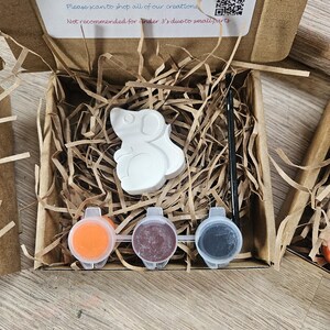 May include: A DIY paint kit in a cardboard box, featuring a white mouse-shaped figurine, three paint pots in orange, maroon, and gray, and a paintbrush. The box contains shredded paper for protection. Instructions are printed on the box.