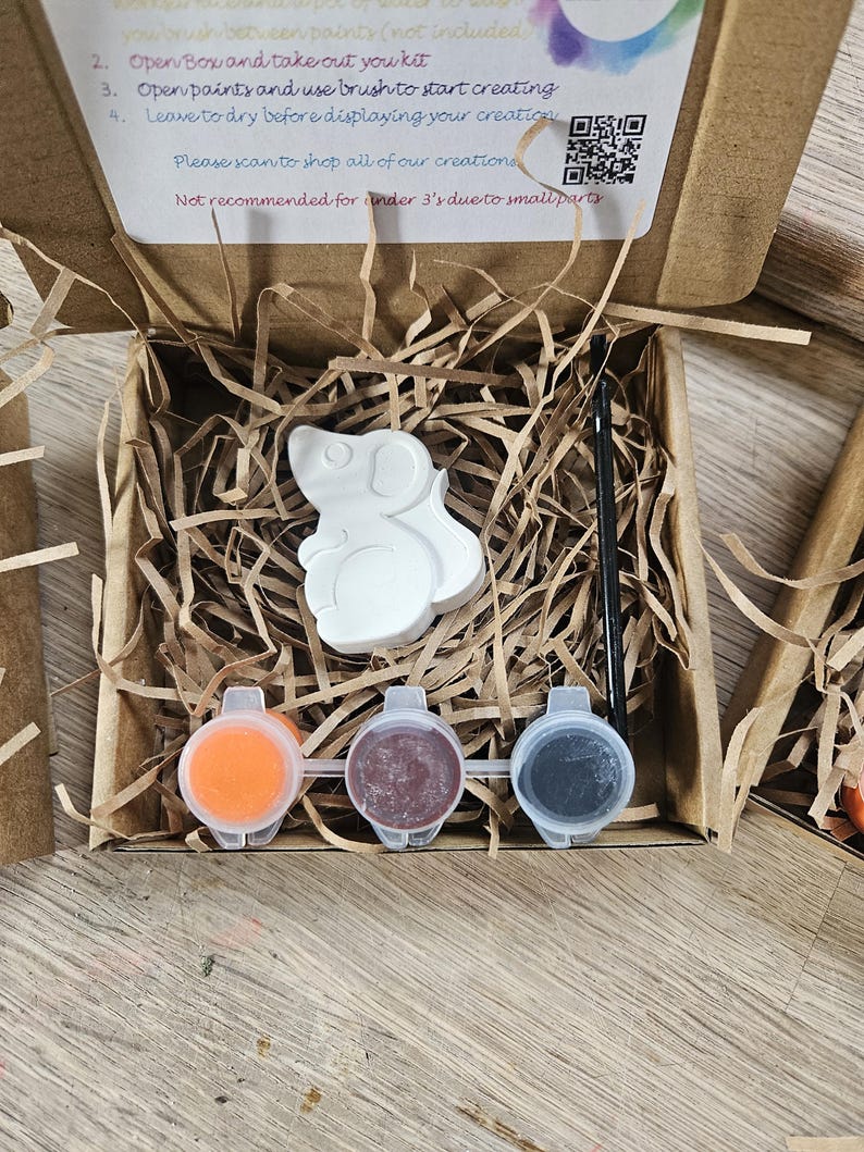 May include: A DIY paint kit in a cardboard box, featuring a white mouse-shaped figurine, three paint pots in orange, maroon, and gray, and a paintbrush. The box contains shredded paper for protection. Instructions are printed on the box.