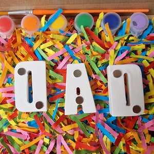Op de afbeelding: Een knutselset voor kinderen om de letters "DAD" te beschilderen met kleurrijke verf. De set bevat een kwast, zes kleine verfpotjes in rood, oranje, geel, groen, blauw en paars, en drie witte houten letters. De letters zijn omringd door kleurrijk papier.