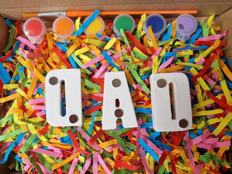 Op de afbeelding: Een knutselset voor kinderen om de letters "DAD" te beschilderen met kleurrijke verf. De set bevat een kwast, zes kleine verfpotjes in rood, oranje, geel, groen, blauw en paars, en drie witte houten letters. De letters zijn omringd door kleurrijk papier.
