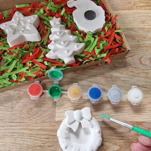 May include: A set of five white ceramic Christmas ornaments, including a star, a tree, a wreath, a Santa Claus, and a bell. The ornaments are in a box with red and green shredded paper. There are six small paint pots with red, green, yellow, blue, gray, and white paint. A green paintbrush is shown in the image.