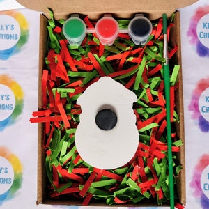 May include: A white ceramic snowman ornament with a black circle for a face, ready to be painted. The ornament is in a box with red and green shredded paper, three small paint pots, and a paintbrush. The paint pots are green, red, and black. The box is labeled "Kelly's Creations".