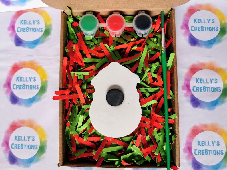 May include: A white ceramic snowman ornament with a black circle for a face, ready to be painted. The ornament is in a box with red and green shredded paper, three small paint pots, and a paintbrush. The paint pots are green, red, and black. The box is labeled "Kelly's Creations".