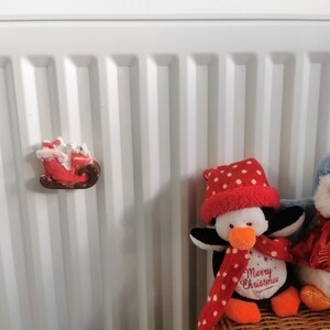 May include: A red and brown Santa Claus sleigh magnet with a white figure, and two penguin stuffed animals. One penguin is black and white with a red scarf and hat, and the other is blue with a red scarf. The penguin has the text "Merry Christmas".