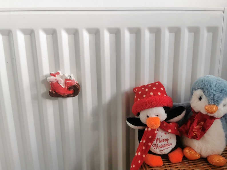 May include: A red and brown Santa Claus sleigh magnet with a white figure, and two penguin stuffed animals. One penguin is black and white with a red scarf and hat, and the other is blue with a red scarf. The penguin has the text "Merry Christmas".
