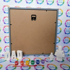 May include: A gray picture frame with a white background, a set of white letters spelling "DAD", a small white figurine of a person, a larger white figurine of a person, and six small paint pots with red, orange, yellow, green, blue, and purple paint. A paintbrush is lying on the table in front of the paint pots.