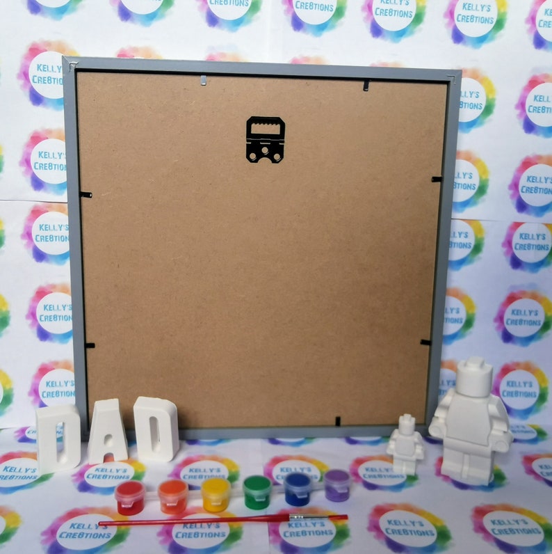 May include: A gray picture frame with a white background, a set of white letters spelling "DAD", a small white figurine of a person, a larger white figurine of a person, and six small paint pots with red, orange, yellow, green, blue, and purple paint. A paintbrush is lying on the table in front of the paint pots.