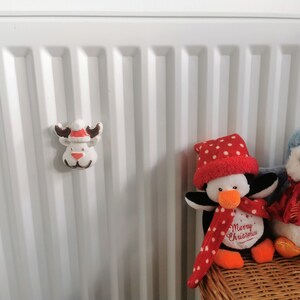 May include: A white reindeer Christmas ornament with a red and white Santa hat, brown antlers, and a red nose is attached to a white radiator. Two plush penguins are in the background.