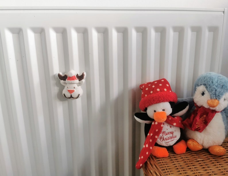 May include: A white reindeer Christmas ornament with a red and white Santa hat, brown antlers, and a red nose is attached to a white radiator. Two plush penguins are in the background.