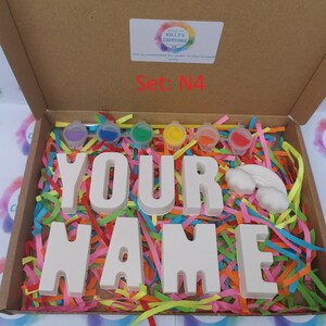 May include: A craft kit in a cardboard box, featuring the words "YOUR NAME" in large, white letters. The box contains paint pots in various colors, a rainbow, and colorful paper strips. The text "Set: N4" is visible.