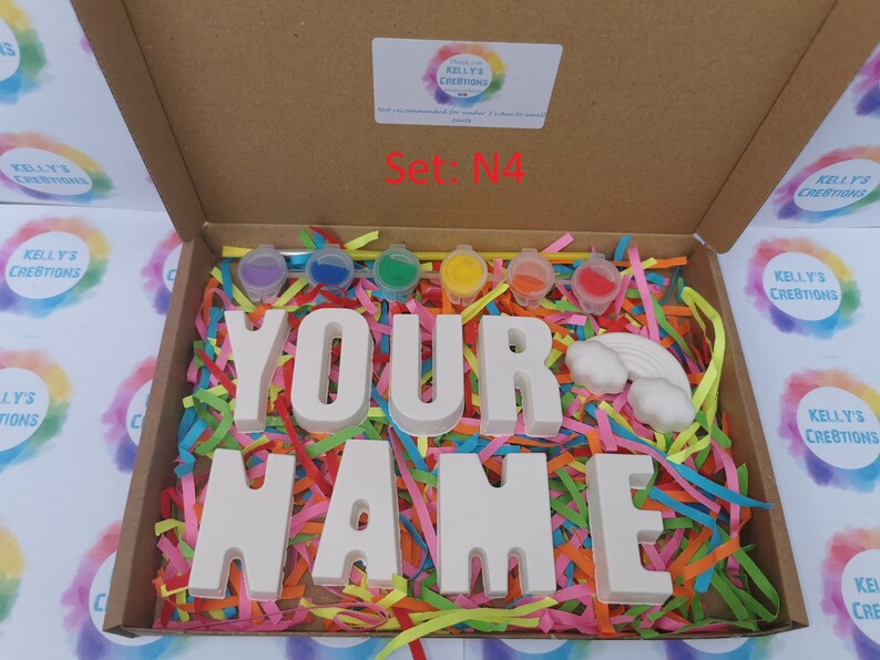 May include: A craft kit in a cardboard box, featuring the words "YOUR NAME" in large, white letters. The box contains paint pots in various colors, a rainbow, and colorful paper strips. The text "Set: N4" is visible.