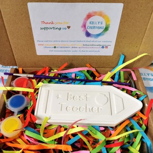May include: A white ceramic pencil shaped plaque with the words "Best Teacher" on it. The plaque is surrounded by colorful shredded paper and comes with a paintbrush and three small pots of paint in red, blue, and yellow.