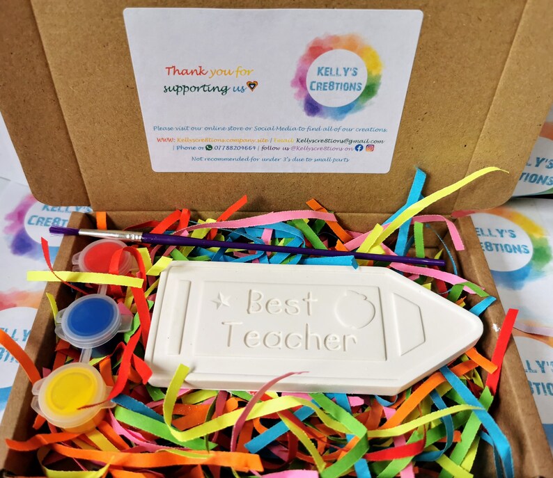 May include: A white ceramic pencil shaped plaque with the words "Best Teacher" on it. The plaque is surrounded by colorful shredded paper and comes with a paintbrush and three small pots of paint in red, blue, and yellow.