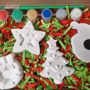 May include: A Christmas craft kit with five white ceramic ornaments: a star, a tree, a wreath, a bell, and a snowman. The kit includes six paint pots in red, green, yellow, blue, silver, and white, and a paintbrush.