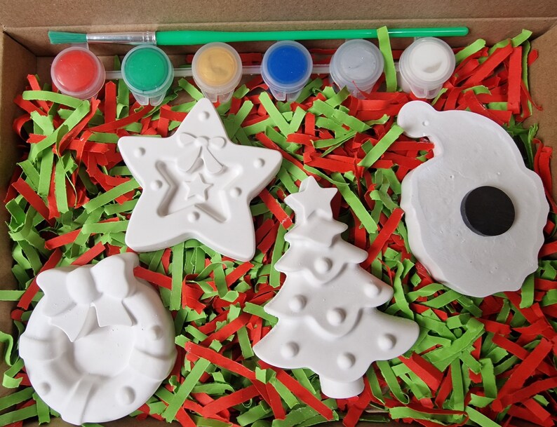 May include: A Christmas craft kit with five white ceramic ornaments: a star, a tree, a wreath, a bell, and a snowman. The kit includes six paint pots in red, green, yellow, blue, silver, and white, and a paintbrush.