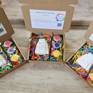 May include: Three open cardboard boxes containing a white plaster sweatshirt figurine embossed with "Class of 2026," a small black paintbrush, and three circular paint pots in red, blue, and yellow, all nestled in colorful shredded paper filler.