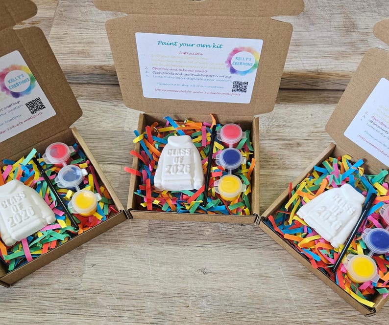 May include: Three open cardboard boxes containing a white plaster sweatshirt figurine embossed with "Class of 2026," a small black paintbrush, and three circular paint pots in red, blue, and yellow, all nestled in colorful shredded paper filler.