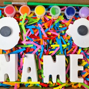 May include: A craft kit with white ceramic letters spelling "NAME" and two white ceramic circles with black magnets. The kit includes five small pots of paint in red, orange, yellow, green, and blue, and a paintbrush. The letters and circles are surrounded by colorful shredded paper.
