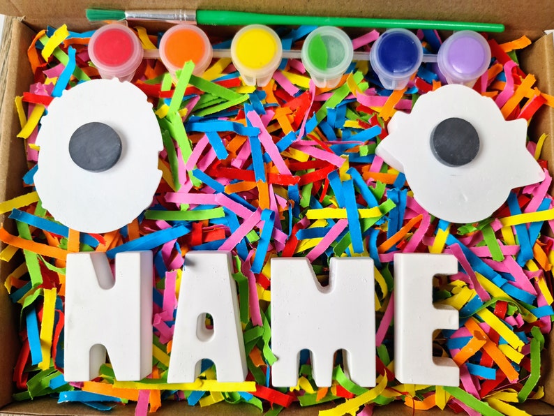 May include: A craft kit with white ceramic letters spelling "NAME" and two white ceramic circles with black magnets. The kit includes five small pots of paint in red, orange, yellow, green, and blue, and a paintbrush. The letters and circles are surrounded by colorful shredded paper.