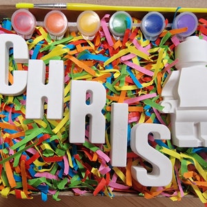 May include: A white ceramic figurine of a person, along with the letters C, H, R, I, S, and T, in white ceramic, all in a box filled with colorful shredded paper. There are six small containers of paint and a paintbrush in the box.