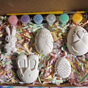 May include: A set of five ceramic Easter-themed figurines in a box with colorful tissue paper. The figurines include a bunny, a bunny with ears, an egg with the words "Happy Easter", an egg with a bird design, and an egg with a zig-zag pattern. The set also includes five small bottles of paint and a paintbrush.