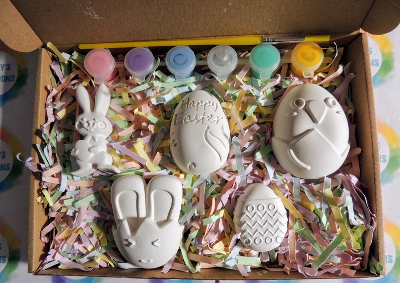 May include: A set of five ceramic Easter-themed figurines in a box with colorful tissue paper. The figurines include a bunny, a bunny with ears, an egg with the words "Happy Easter", an egg with a bird design, and an egg with a zig-zag pattern. The set also includes five small bottles of paint and a paintbrush.