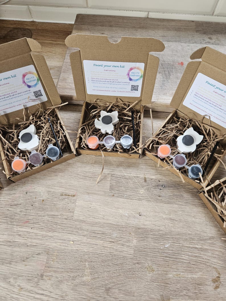 May include: Several craft kits in open cardboard boxes. Each kit contains a white ceramic flower-shaped magnet, paint pots in orange, purple, and silver, a paintbrush, and instructions. The boxes are filled with shredded paper for protection. The text on the box reads "Paint your own kit".