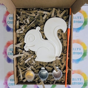 K&ouml;nnte beinhalten: Eine wei&szlig;e Keramik-Eichh&ouml;rnchenfigur in einem Karton mit Farbt&ouml;pfen und einem Pinsel. Der Karton ist mit braunen Verpackungschips gef&uuml;llt. Die Farbt&ouml;pfe sind gold, silber und braun. Der Pinsel ist orange.