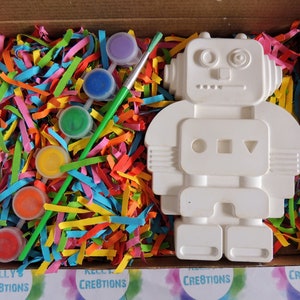 May include: A white ceramic robot figurine with paint-by-number sections, a paintbrush, and six small paint pots in various colors. The robot is in a cardboard box with colorful shredded paper.