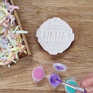 May include: A white ceramic round ornament with the text "Best Nana Ever" in a decorative font. The ornament is surrounded by small hearts and arrows. Three small paint pots with pink, purple, and teal paint are on a wooden surface. A paintbrush is dipping into the purple paint.