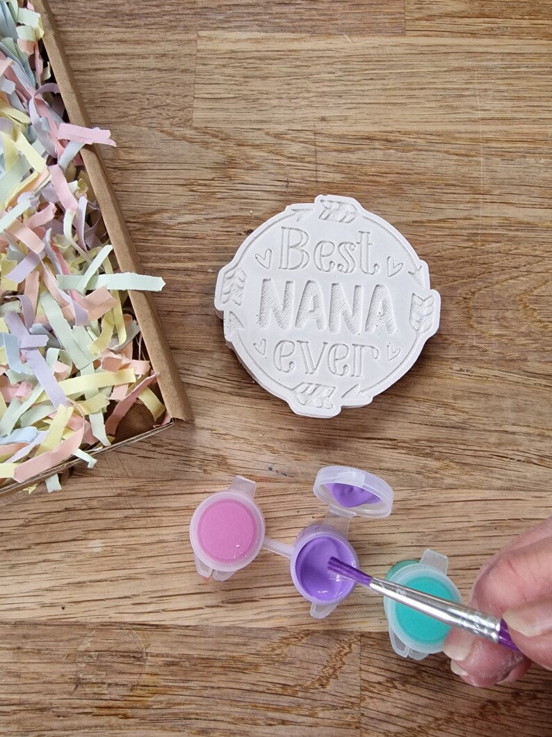May include: A white ceramic round ornament with the text "Best Nana Ever" in a decorative font. The ornament is surrounded by small hearts and arrows. Three small paint pots with pink, purple, and teal paint are on a wooden surface. A paintbrush is dipping into the purple paint.