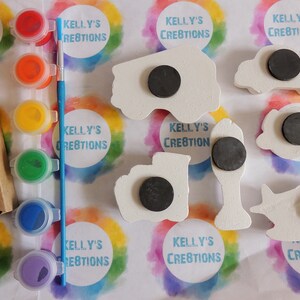 May include: A set of five wooden vehicle shapes with magnetic backs, ready to be painted. The set includes a fire truck, a car, a bus, a truck, and a plane. The vehicles are white and have black magnetic circles on the back. The set also includes a set of four paint pots with red, yellow, green, and blue paint, and a blue paintbrush.