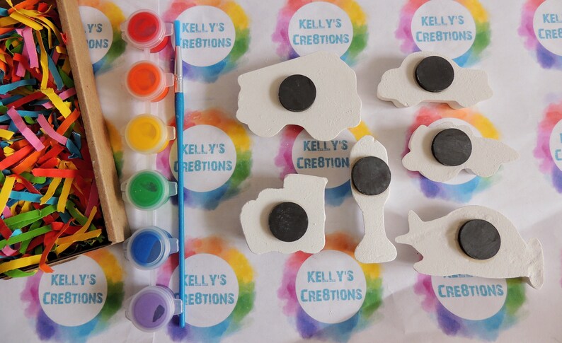 May include: A set of five wooden vehicle shapes with magnetic backs, ready to be painted. The set includes a fire truck, a car, a bus, a truck, and a plane. The vehicles are white and have black magnetic circles on the back. The set also includes a set of four paint pots with red, yellow, green, and blue paint, and a blue paintbrush.