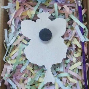 May include: A white ceramic fairy figurine with a magnet on the back. The fairy has wings and a crown. The figurine is surrounded by colorful shredded paper and three small paint pots with lids. A purple paintbrush is also included.