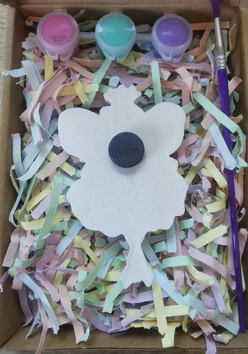 May include: A white ceramic fairy figurine with a magnet on the back. The fairy has wings and a crown. The figurine is surrounded by colorful shredded paper and three small paint pots with lids. A purple paintbrush is also included.