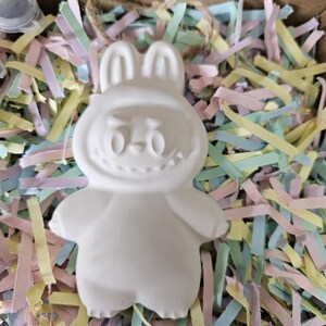 Peut inclure: Une figurine de lapin en c&eacute;ramique blanche avec un visage souriant, des oreilles et des bras, pos&eacute;e sur du papier d&eacute;chiquet&eacute; color&eacute;. Le lapin est dans une bo&icirc;te en carton, pr&ecirc;t &agrave; &ecirc;tre peint. Un petit pot de peinture est visible.