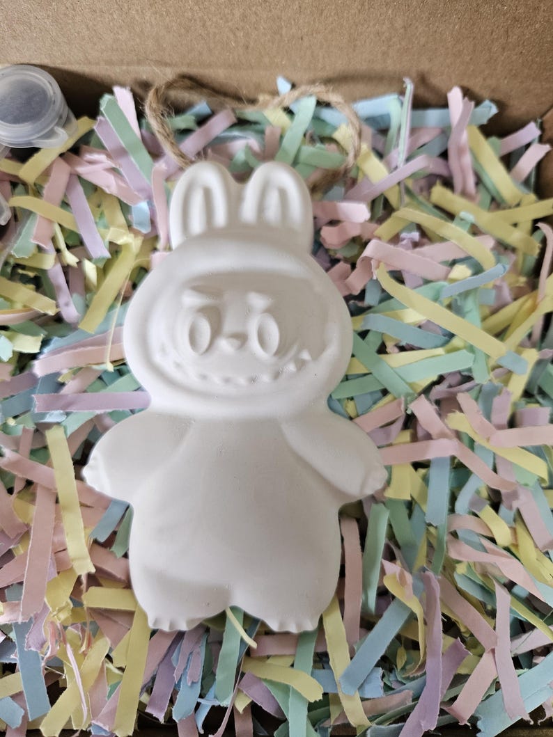 Peut inclure: Une figurine de lapin en c&eacute;ramique blanche avec un visage souriant, des oreilles et des bras, pos&eacute;e sur du papier d&eacute;chiquet&eacute; color&eacute;. Le lapin est dans une bo&icirc;te en carton, pr&ecirc;t &agrave; &ecirc;tre peint. Un petit pot de peinture est visible.