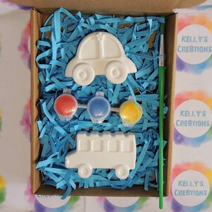 Puede incluir: Un kit de manualidades en una caja de cart&oacute;n, con un coche y un autob&uacute;s escolar blancos, botes de pintura roja, azul y amarilla, y un pincel verde. La caja est&aacute; llena de papel triturado azul. El texto "Kelly's Creations" es visible.