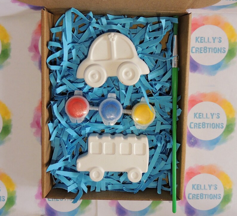 Puede incluir: Un kit de manualidades en una caja de cart&oacute;n, con un coche y un autob&uacute;s escolar blancos, botes de pintura roja, azul y amarilla, y un pincel verde. La caja est&aacute; llena de papel triturado azul. El texto "Kelly's Creations" es visible.