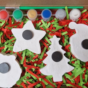 May include: A Christmas craft kit with five white ceramic ornaments, a paintbrush, and six paint pots. The ornaments are shaped like a star, a tree, a wreath, a bell, and a stocking. The paint pots are red, green, yellow, blue, white, and silver.