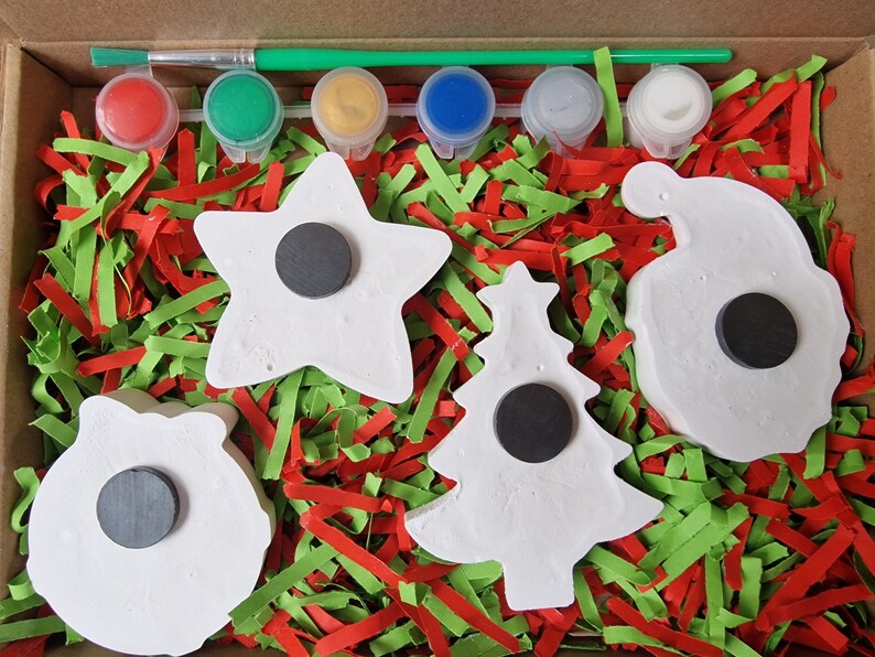 May include: A Christmas craft kit with five white ceramic ornaments, a paintbrush, and six paint pots. The ornaments are shaped like a star, a tree, a wreath, a bell, and a stocking. The paint pots are red, green, yellow, blue, white, and silver.