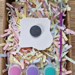 May include: A craft kit with a white ceramic tile, a black circle magnet, three small paint pots in pink, purple, and teal, and a purple paintbrush. The kit is packaged in a cardboard box with shredded pastel paper.