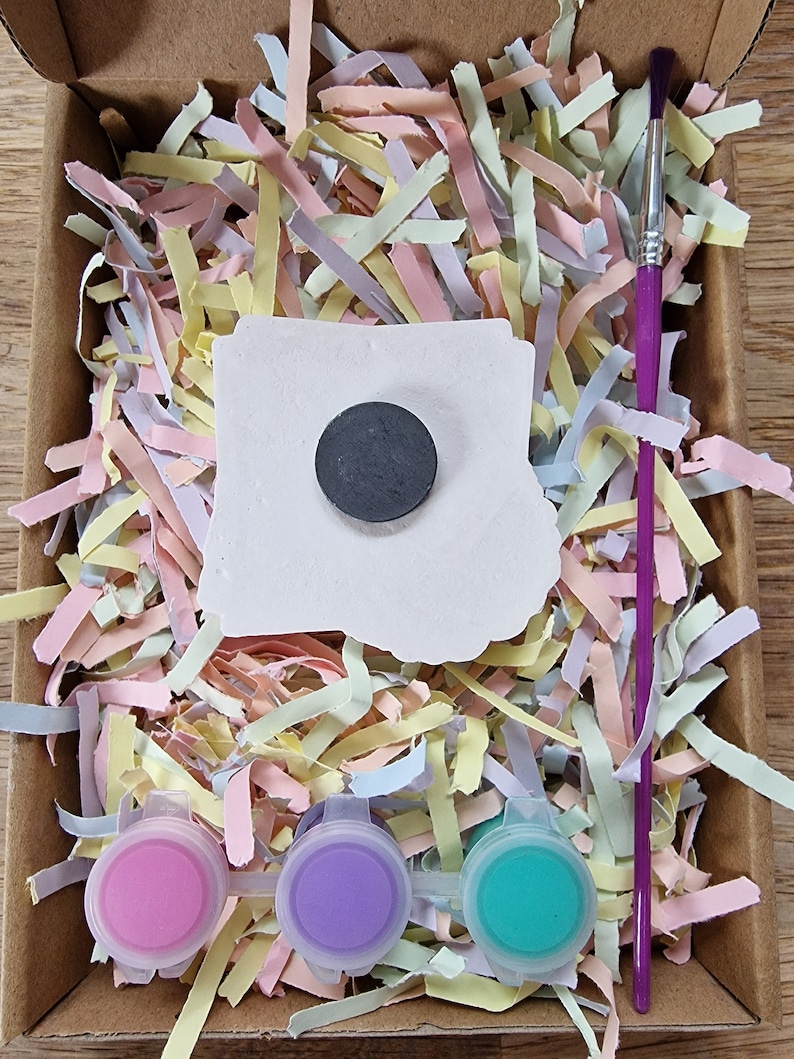 May include: A craft kit with a white ceramic tile, a black circle magnet, three small paint pots in pink, purple, and teal, and a purple paintbrush. The kit is packaged in a cardboard box with shredded pastel paper.