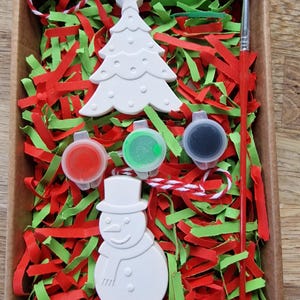 white plaster bauble with paints and brush in a box with green and red shred.