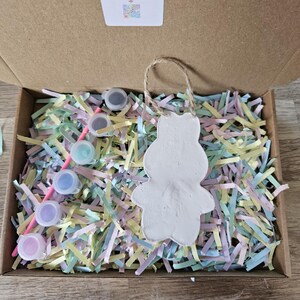 Peut inclure: Un kit de bricolage dans une bo&icirc;te en carton, comprenant un ornement de lapin en c&eacute;ramique blanche avec une boucle en ficelle. La bo&icirc;te contient du papier d&eacute;chiquet&eacute; color&eacute;, de petits pots de peinture et un pinceau. Le kit est de Kelly's Creations.