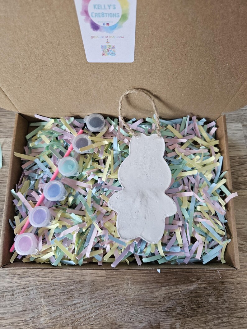 Peut inclure: Un kit de bricolage dans une bo&icirc;te en carton, comprenant un ornement de lapin en c&eacute;ramique blanche avec une boucle en ficelle. La bo&icirc;te contient du papier d&eacute;chiquet&eacute; color&eacute;, de petits pots de peinture et un pinceau. Le kit est de Kelly's Creations.